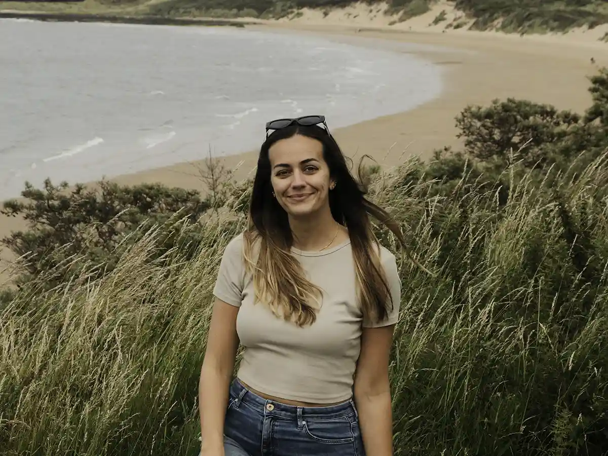 about-me-beach-portrait Lucy smiling at the camera with the curved shoreline of Gullane Beach in the background.